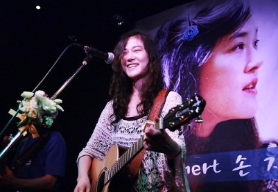 손지연이-꽃으로-장식된-스탠드-마이크-앞에-기타를-메고-서서-웃고-있는-사진