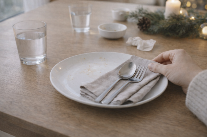 연말 식사를 마친 뒤 깨끗하게 정리된 테이블 위에 접시와 수저가 놓여 있고, 한 손이 접시를 정리하고 있는 장면