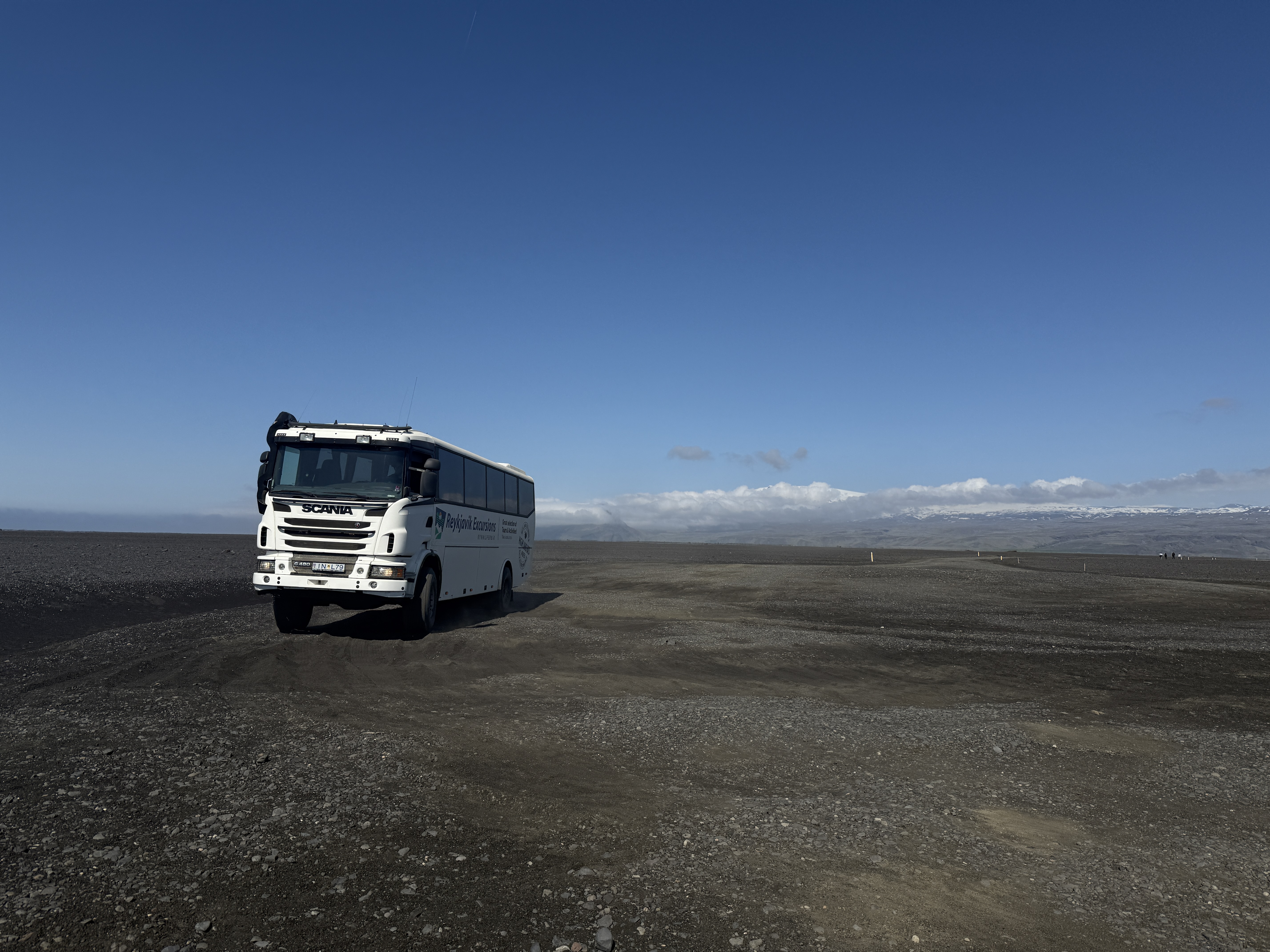 검은 화산재 지형 위를 달리는 하얀색 셔틀버스의 모습입니다. 주변은 완전히 황무지이며, 배경에는 푸른 하늘과 멀리 눈 덮인 산맥과 구름이 있습니다.