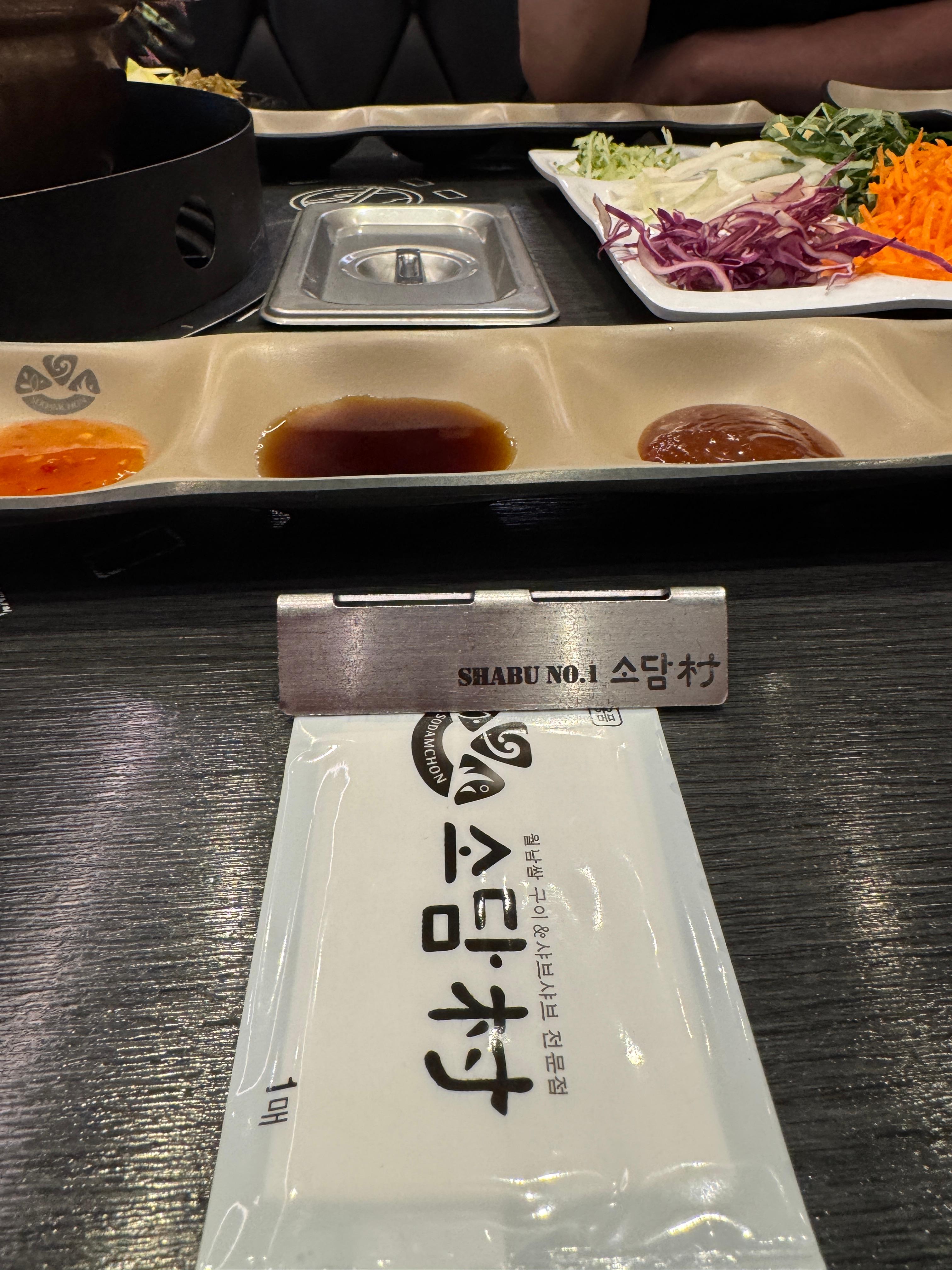 소담촌 김포한강신도시점 샤브샤브 매장의 기본 테이블 세팅. 소스 3종이 담긴 접시와 월남쌈용 채소, 그리고 매장명이 새겨진 물수건과 금속 명패가 보인다. 정갈한 상차림이 샤브샤브 전문점의 분위기를 잘 보여준다.