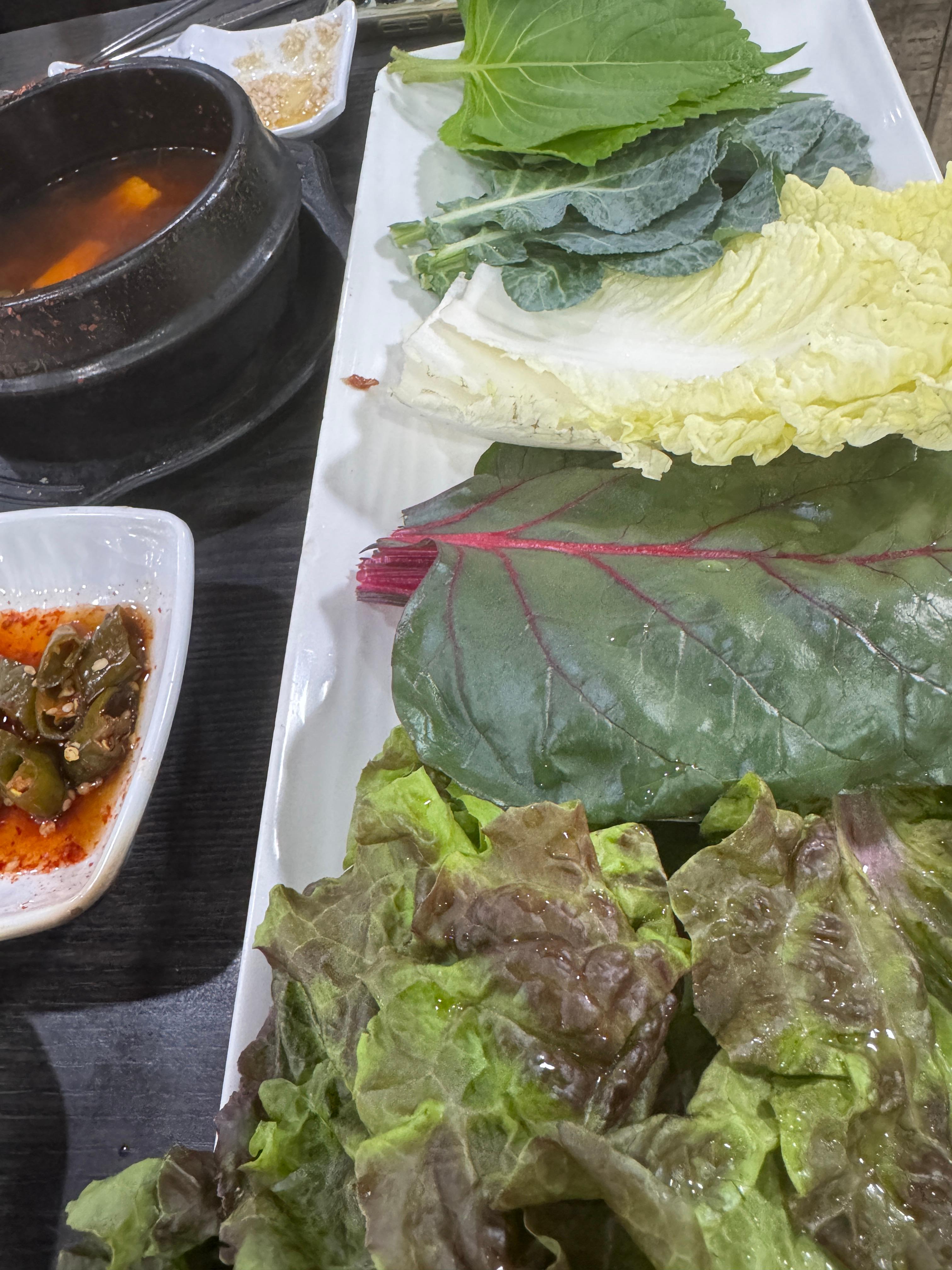 상추&amp;#44; 깻잎&amp;#44; 케일&amp;#44; 청경채&amp;#44; 적근대 등 다양한 쌈채소가 정갈하게 담긴 접시. 옆에 된장찌개와 반찬도 보인다. 