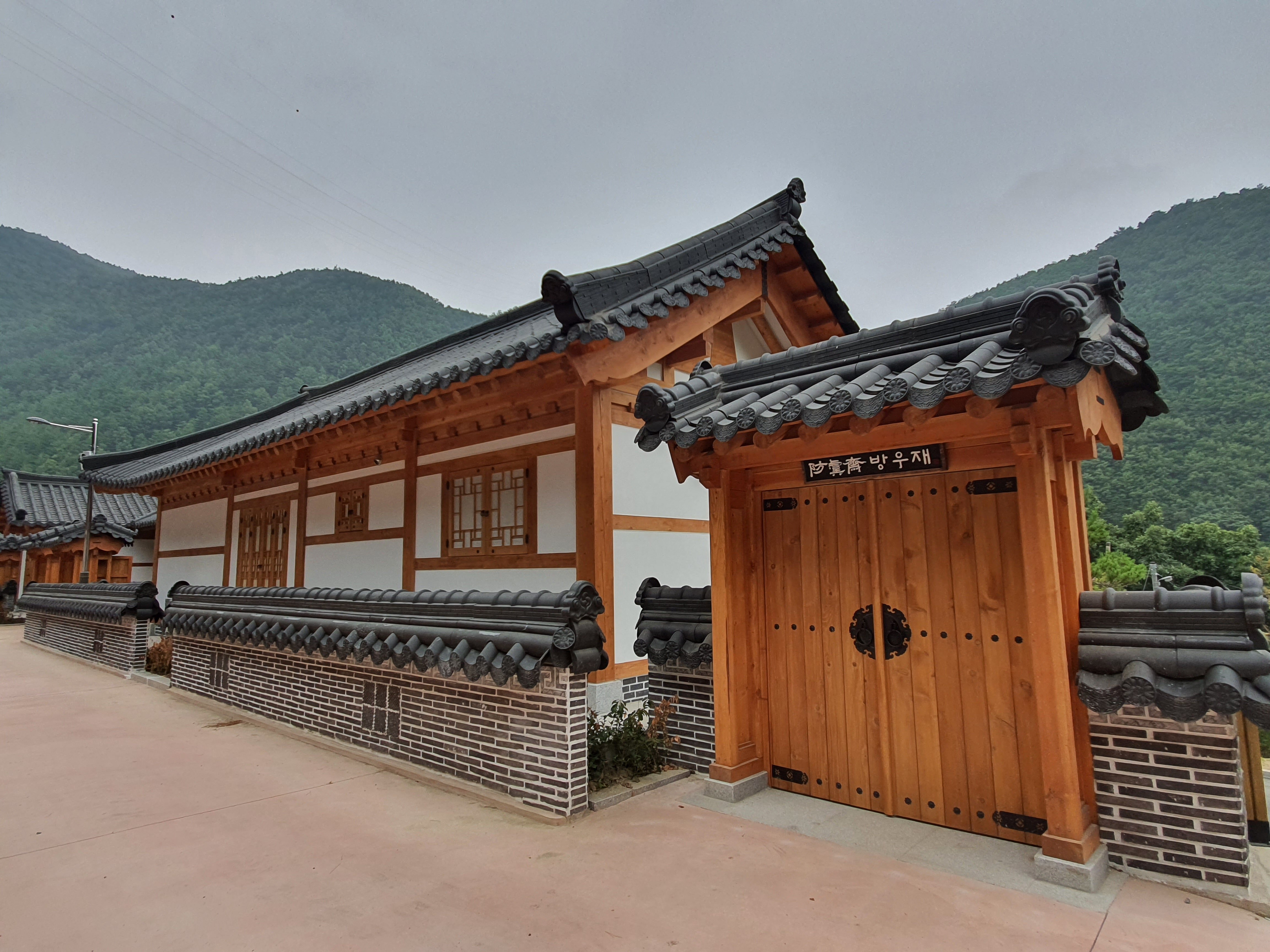 산자락 아래 전통 기와지붕과 나무 기둥이 조화로운 한옥 숙소가 자리 잡고 있으며 주위로 초록빛 나무들이 감싸고 있는 평화로운 모습