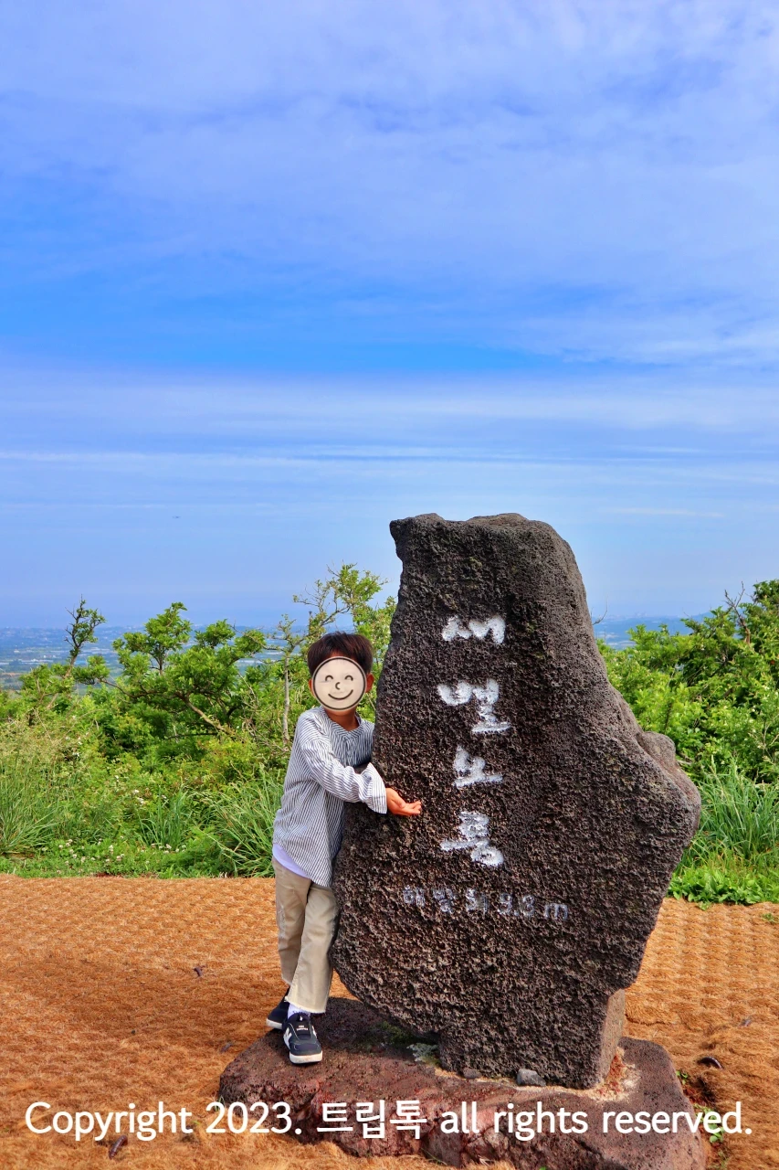 하늘하애 세로로 길에생긴 동에 '새별오름'이라고 하얀글씨가 새겨져있고 그옆에 한 아이가 서있다