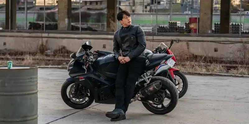 어느 야외를 배경으로 오토바이에 엉덩이를 댄 채 검은색으로 통일한 가죽자켓, 바지로 오른쪽을 바라보고 있는 두영