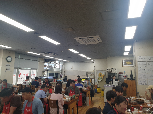 짱구네 내부 사람들로 가득차있다. 바닥은 우드 느낌 벽면은 사무실 같은 처리 위에는 삼열로 조명이 있다.