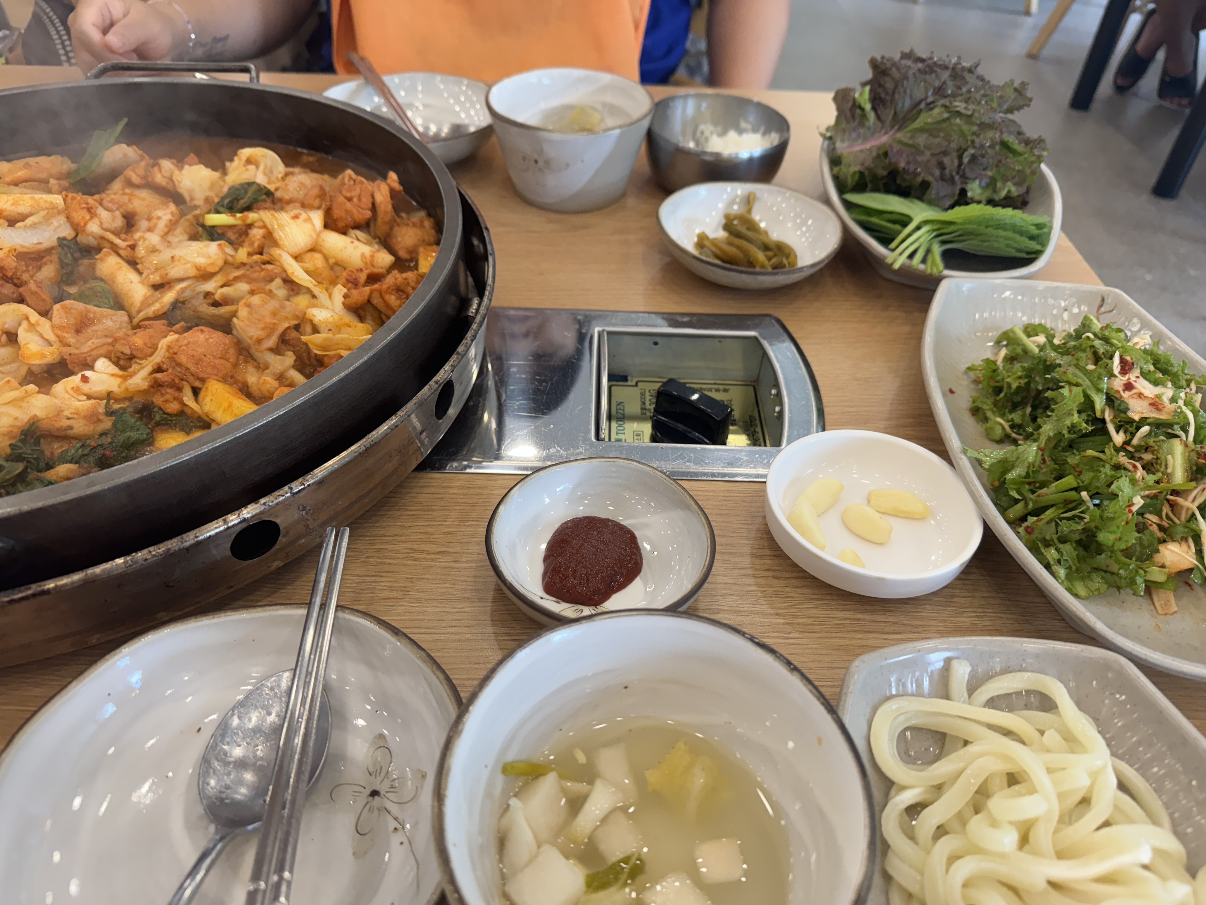 닭갈비와 함께 상에 차려진 반찬들. 상추&amp;#44; 고추&amp;#44; 샐러드&amp;#44; 마늘&amp;#44; 깍두기 국물&amp;#44; 공깃밥&amp;#44; 우동사리가 함께 놓여 있다. 