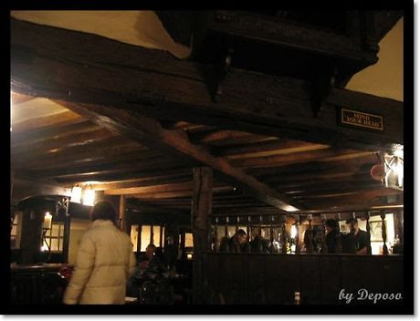 영국 런던 근교의 전통적인 펍 내부. 낮은 천장에는 두꺼운 나무 들보가 드러나 있고, 따뜻한 조명 아래 손님들이 삼삼오오 모여 대화를 나누고 있다. 왼쪽에는 흰색 패딩을 입은 여성이 서 있고, 전체적으로 아늑하고 고풍스러운 분위기를 풍긴다.