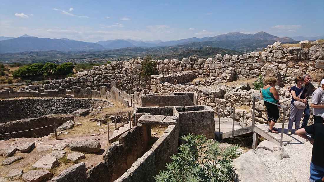 미케네 유적지의 원형 묘지와 여러 고대 석조 구조물, 배경으로 산과 평야가 펼쳐진다.