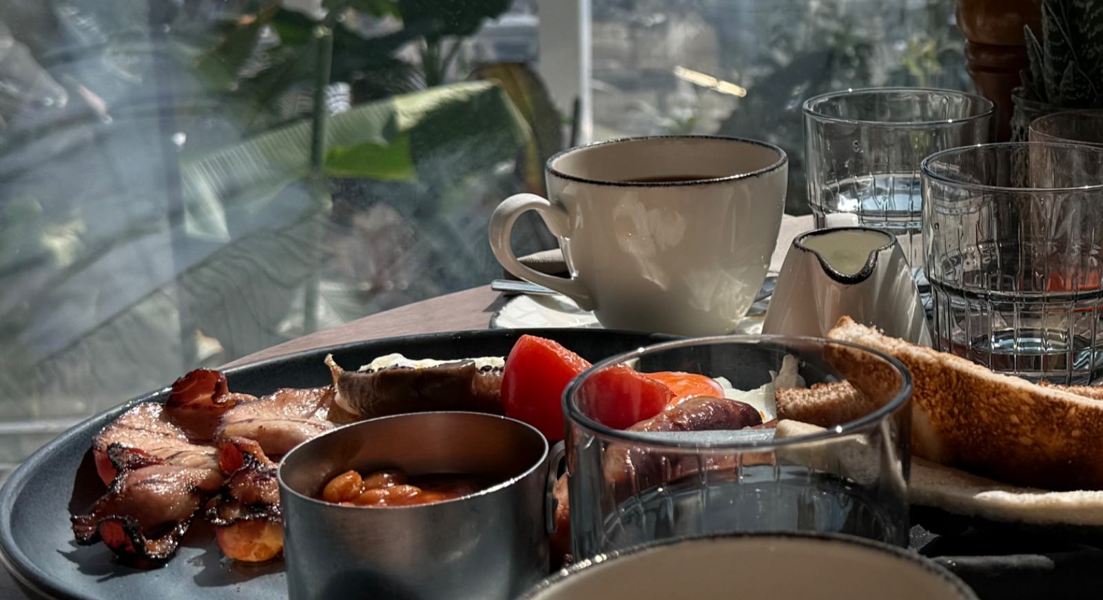 창가 테이블 위에 차려진 잉글리시 브랙퍼스트. 베이컨, 베이크드 빈스, 구운 토마토, 소시지, 토스트와 커피, 유리컵 등이 놓여 있다.