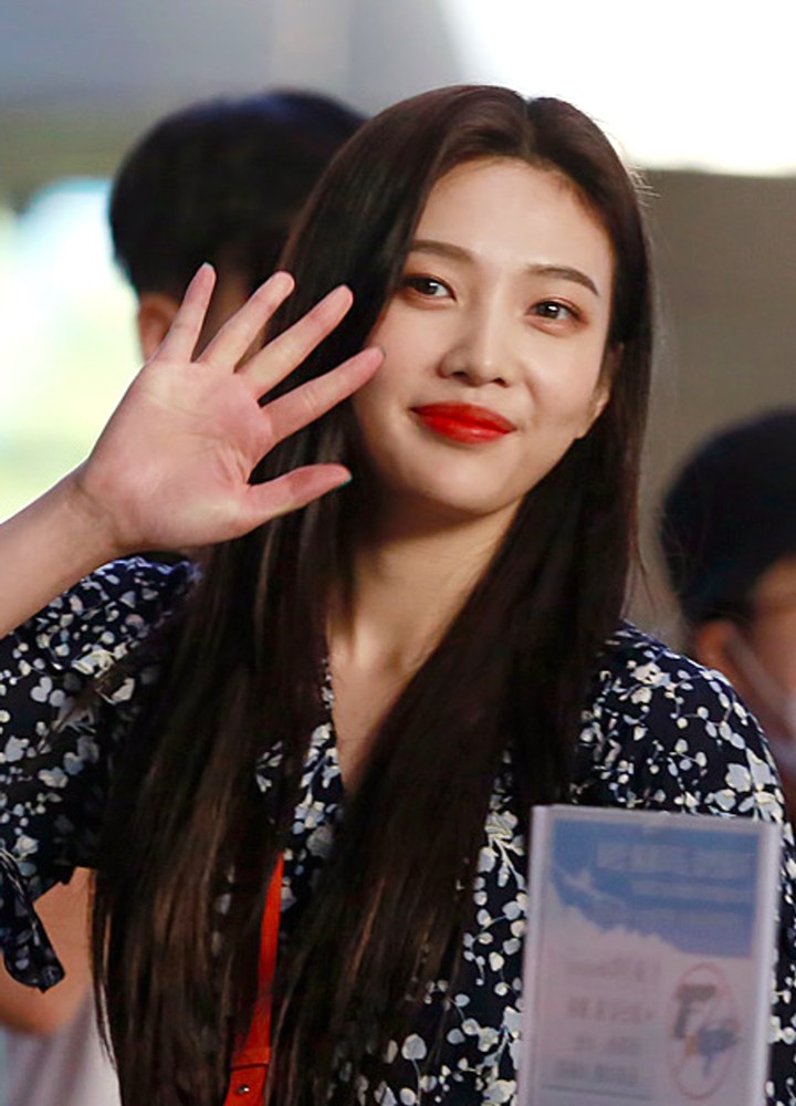 어두운 무늬 옷을 입은 긴 머리 여성이 붉은 립스틱을 바르고 한 손을 들어 인사하는 모습