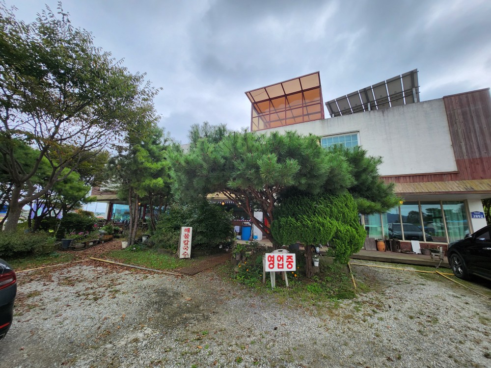 소나무와 아기자기한 조경으로 둘러싸인 좋은만남해물칼국수 식당 건물 외관. '영업중' 푯말이 손님을 반기고 있다