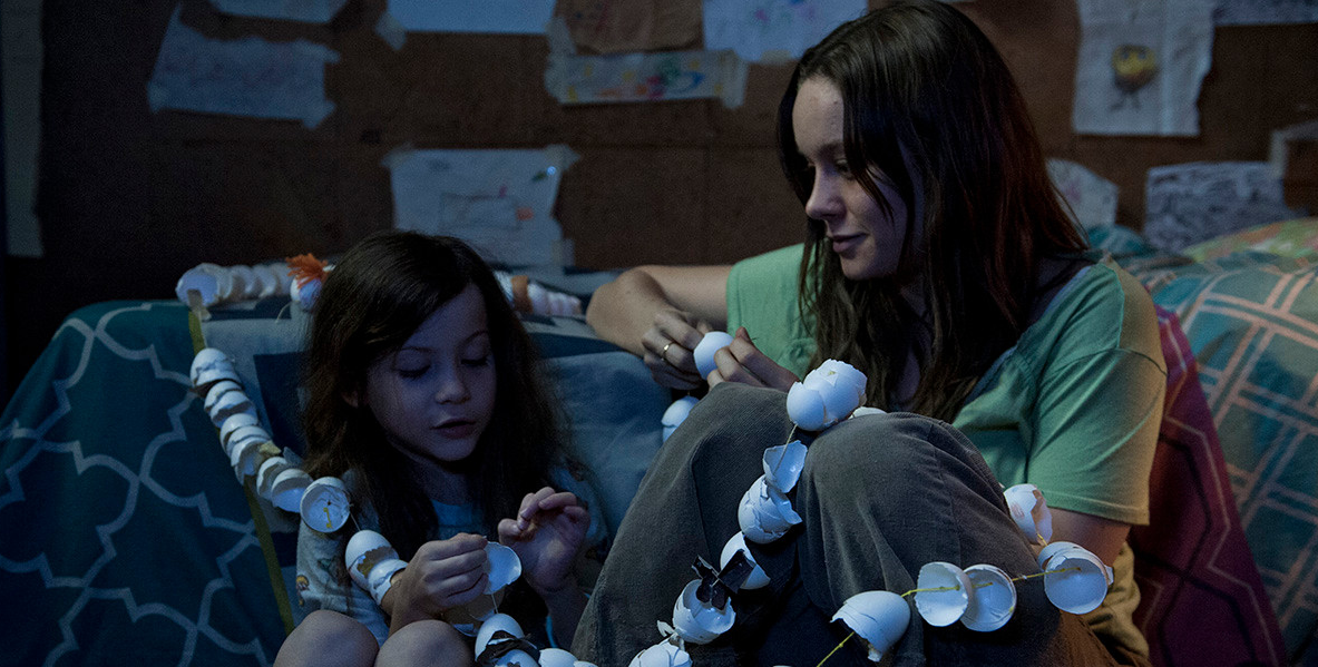 어둑한 방 안에서 잭과 조이가 나란히 앉아 달걀 껍데기로 만든 장식 줄을 함께 엮고 있는 장면, 벽에는 아이가 그린 그림들이 붙어 있다