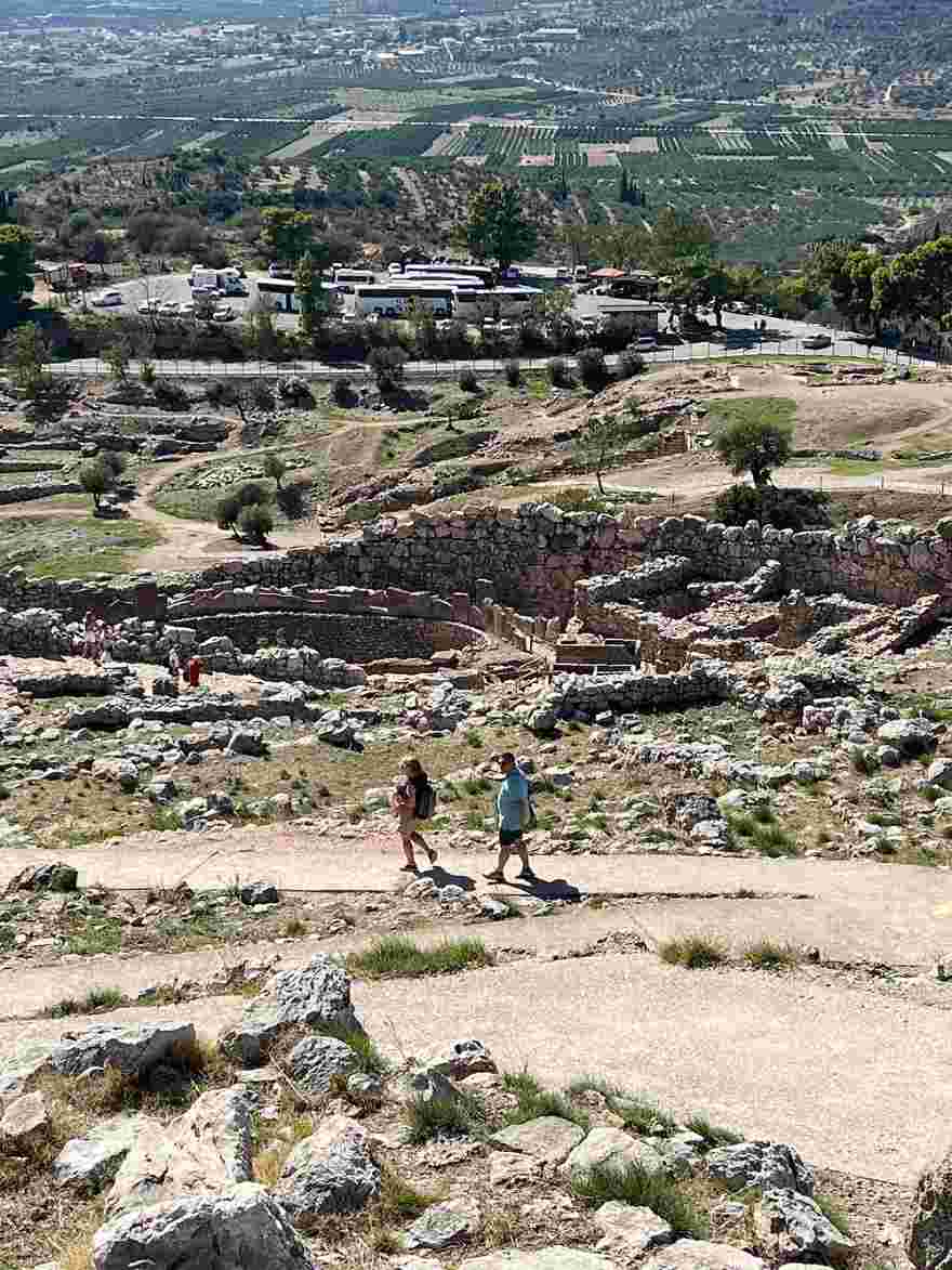 미케네 유적지의 산책로를 따라 걷는 두 사람과 멀리 펼쳐진 성벽, 평야 풍경이 보인다.