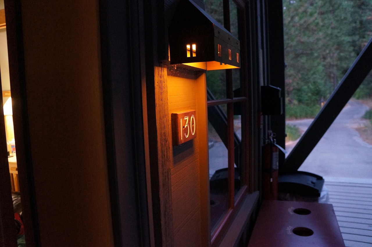 나무 벽면에 부착된 주소판 '130'을 은은하게 비추는 작은 벽등이 중심에 위치한 장면으로, 조명이 들어 있는 미니어처 하우스 형태의 등은 따뜻한 오렌지빛을 내며 포근한 분위기를 자아낸다. 화면 오른쪽으로는 좁은 도로와 주변 숲길이 어슴푸레하게 이어지며 외부의 조용한 자연환경과 대비된다. 왼편 실내에는 은은한 전등과 나무 가구가 보이며, 실내외의 경계가 조화롭게 연결되어 있다. 전반적으로 아늑함과 고요함이 어우러진 전원주택의 초저녁 분위기를 담은 사진.