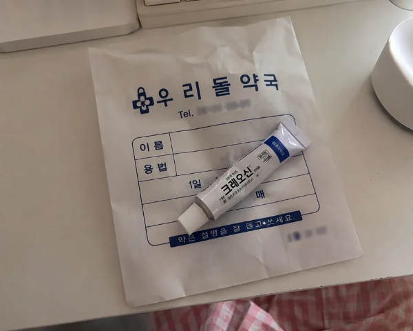 흰색 책상 위에 올려진 우리돌약국 종이 약봉투와 그 위에 툭 놓인 피부과 처방 크레오신 연고 튜브 사진