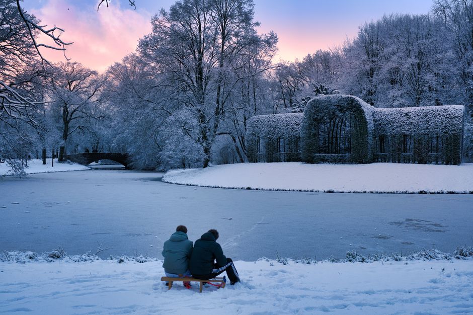 Two people sitting by a frozen lake in a snowy winter landscape at sunset.