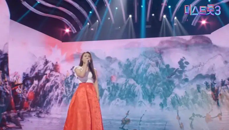 미스트롯3- 흰 반팔티 빨간 긴치마를 입고 오른손으로 마이크를 든 채 긴머리를 늘어뜨리고 노래하는 미스김