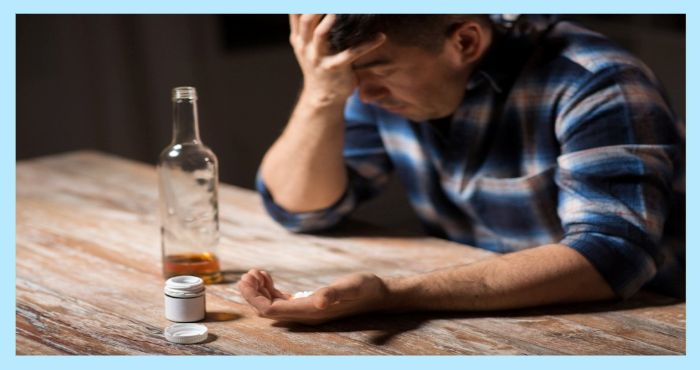 머리를 감싸쥐고 술병과 약병 앞에 앉아 고민하는 남성의 사진. 우울감이나 중독, 자살 위험을 시사하는 상황.