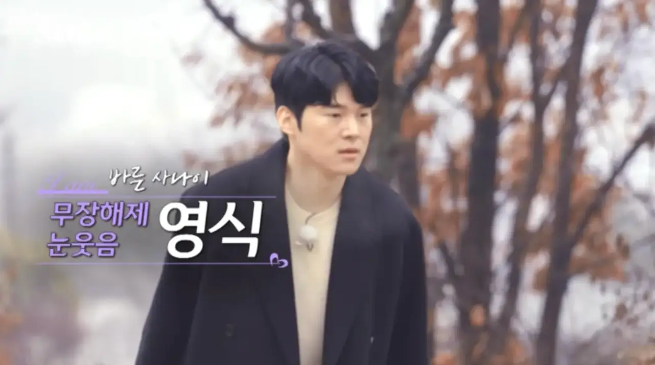 검은코트 안 아이보리색 니트를 입고 걸어가는 영식 좌측 보라색 글씨 무장해제 눈웃음 영식