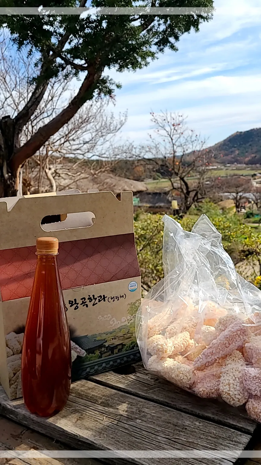 나무테이블 위 왕곡한과박스 한과봉지 수정과한병 파란하늘과 푸른산 소나무 한그루