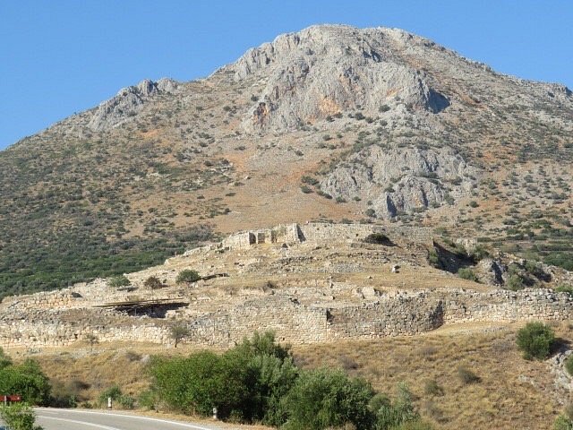 넓은 돌담과 잔디 언덕 위에 자리잡은 고대 미케네 유적지, 뒤쪽에는 커다란 바위산이 우뚝 솟아 있다.