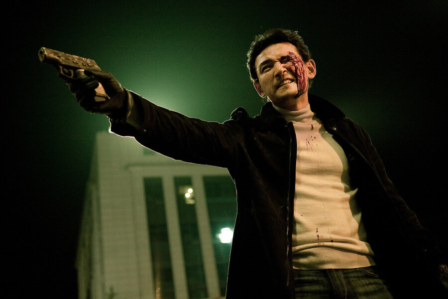 눈 위쪽에 피를 흘리며 얼굴을 찡그린채 한 손으로 총을 겨누고 있는 최철기