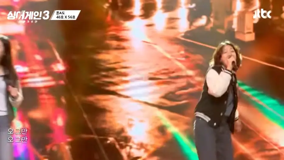 싱어게인 3- 검정색 몸통에 흰색팔부분 점퍼를 입고 단발 곱슬머리상태로 오른손으로 마이크를 쥐고 청바지를 입은채 노래하는 56호