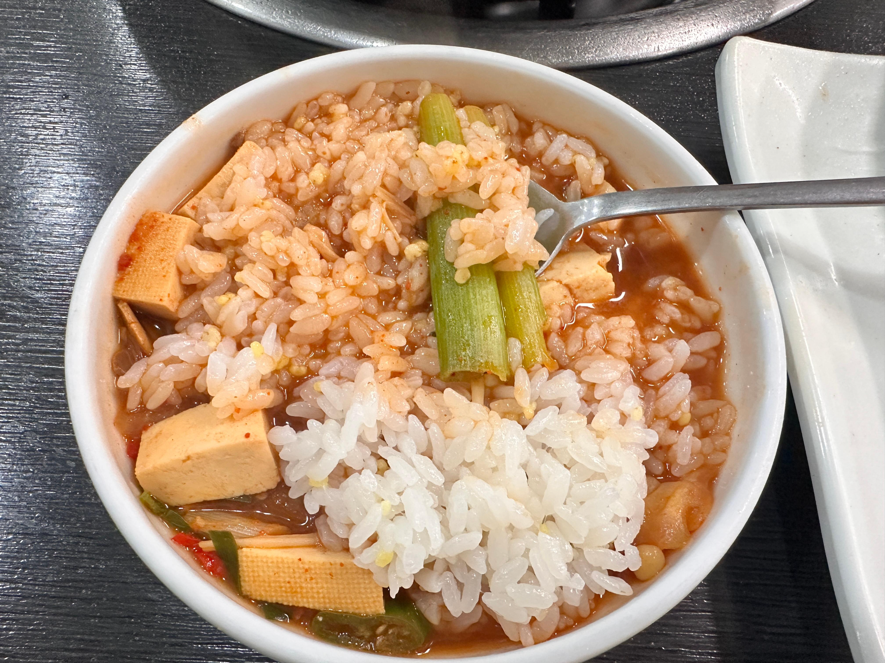 된장찌개 국물과 밥이 섞인 비빔밥이 하얀 공기에 담겨있는 모습. 두부와 채소 조각들이 보임