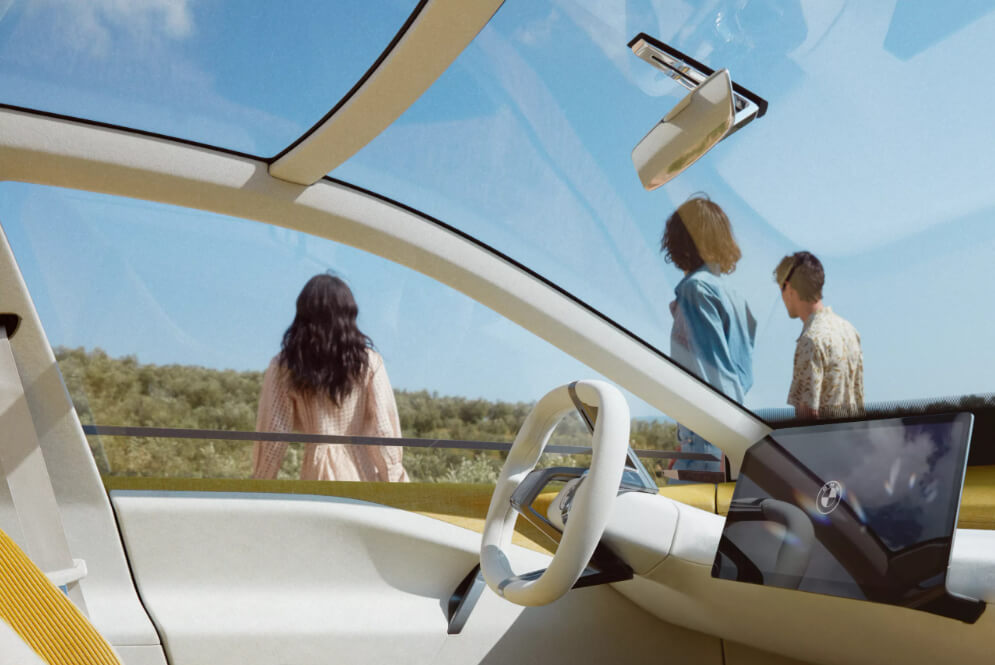 BMW Neue Klasse 전기차의 넓고 미래지향적인 내부가 보입니다. 파노라마 유리 지붕과 심플한 스티어링 휠이 특징적이며, 차량 밖으로는 맑은 날씨에 사람들이 걸어가는 모습이 보입니다.