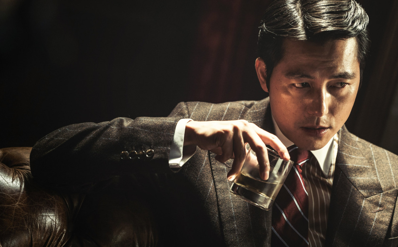 어두운 조명 아래 가죽 소파에 기대어 위스키 잔을 들고 앞을 응시하는 태수의 클로즈업 장면