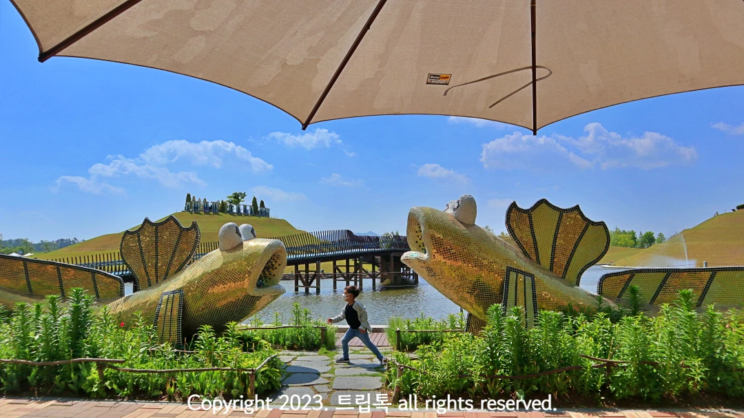 파라솔 아래 호수가 보이고 짱뚱어 조형물이 양쪽에 있고 가운에 한 아이가 뛰고 있다.