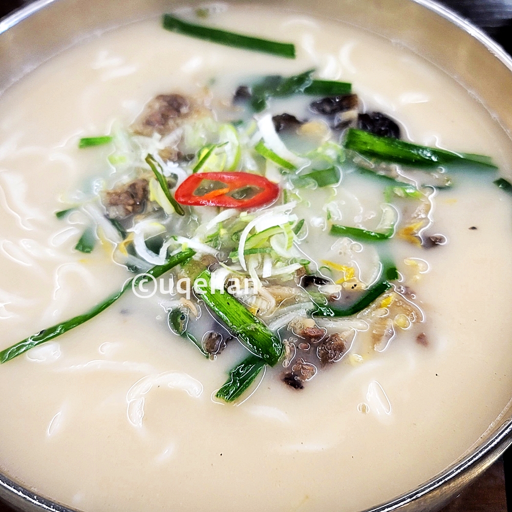 양주 밀곳 칼국수 사진이다. 뽀얀 국물 안에 칼국수 면이 들어가 있고, 부추와 쑥주나물 그리고 목이버섯이 고명으로 올라가 있다. 맨 위에는 빨강고추를 작게 잘라 올려놓았다.