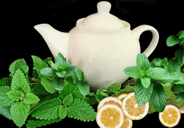 검정 바탕에 하얀 도자기 차주전자가 있고, 앞에는 초록색잎을 가진 레몬밤들과 얇게 저민 레몬 슬라이스들이 있다.