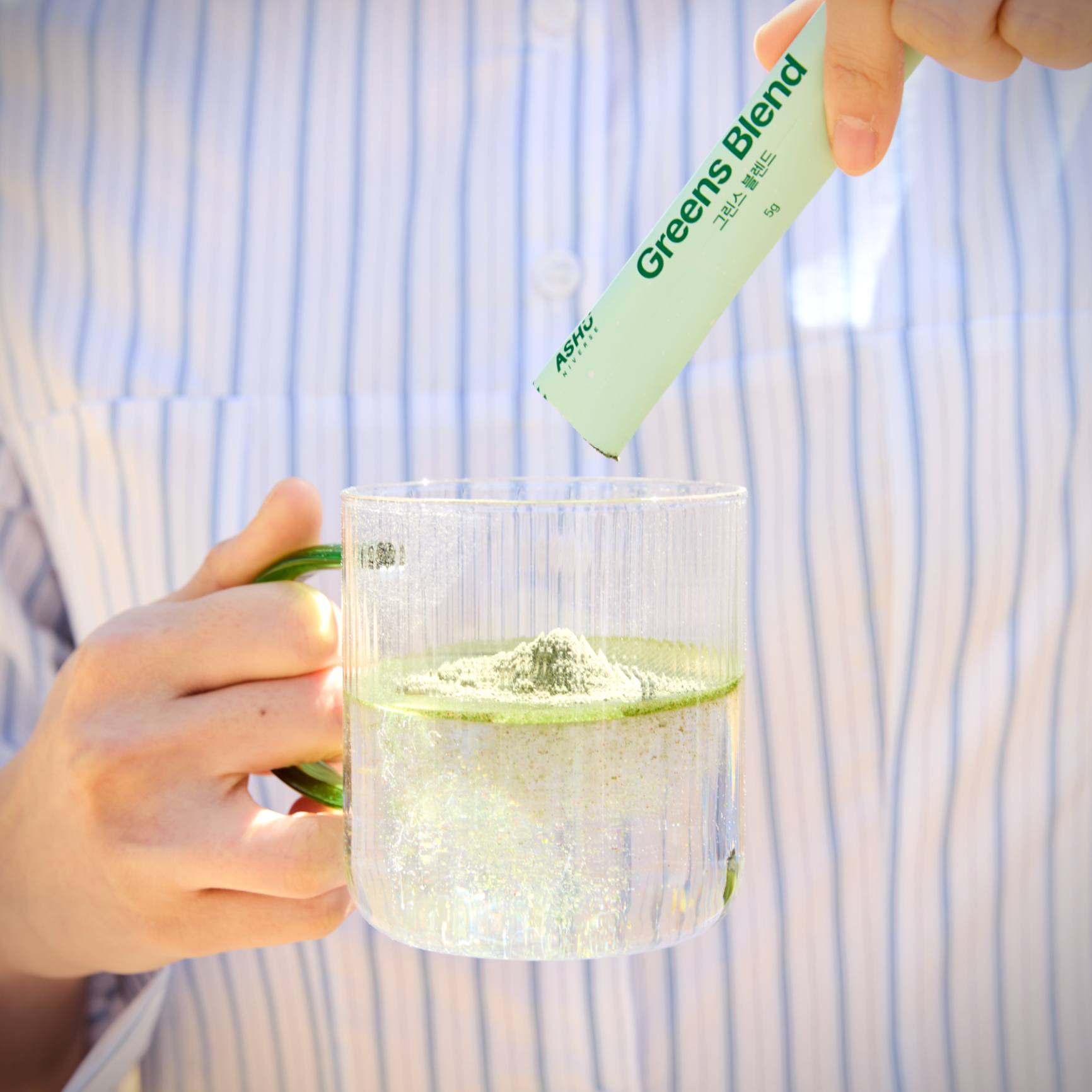 아슈니버스 그린스 블렌드 분말을 물에 타서 섞는 장면의 클로즈업 이미지