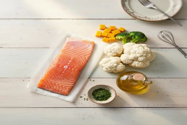 깨끗한 나무 테이블 위에서, 신선한 연어 한 토막, 깨끗이 손질된 콜리플라워 (또는 몇 개의 대체 채소 조각), 올리브 오일 병, 작은 간식용 파슬리 가루 통이 정갈하게 놓여있는 탑다운(Top-down) 뷰 이미지입니다.