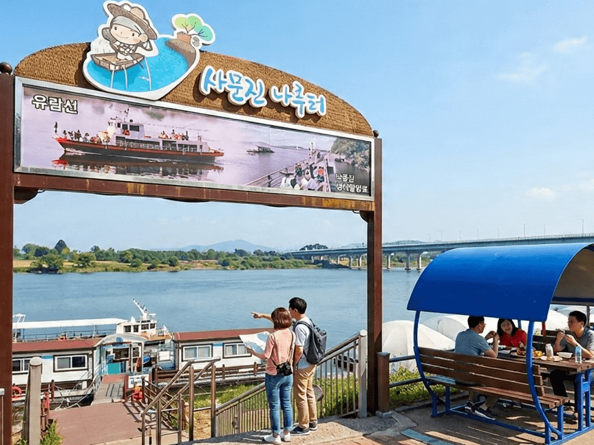대구 달성군 화원유원지 사문진나루터 입구에 유람선 사진과 캐릭터 일러스트가 있는 아치형 안내 조형물이 있고 아래에 낙동강과 유람선 선착장, 강변 벤치가 보이는 맑은 날 전경