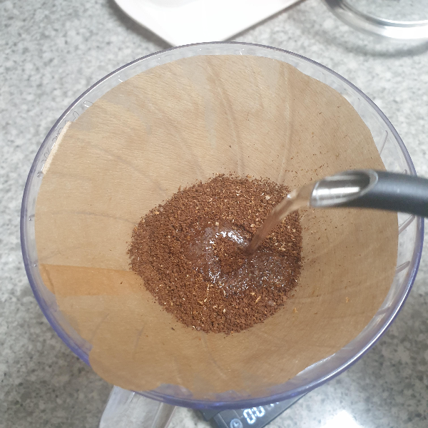 하리오 드리퍼에 황색 필터가 장착되어 있다. 필터 안에는 원두가 있고 드립포트 추출구에서 물이 나오고 있다.