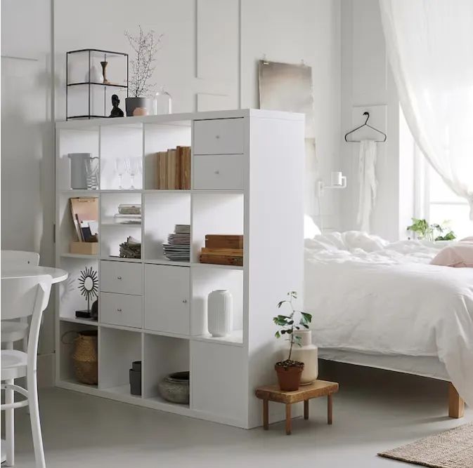 침실-흰색칼락스책상-책과그릇이진열된-서랍또는문이달린칸이있는-책장뒤에흰색침대가있다
