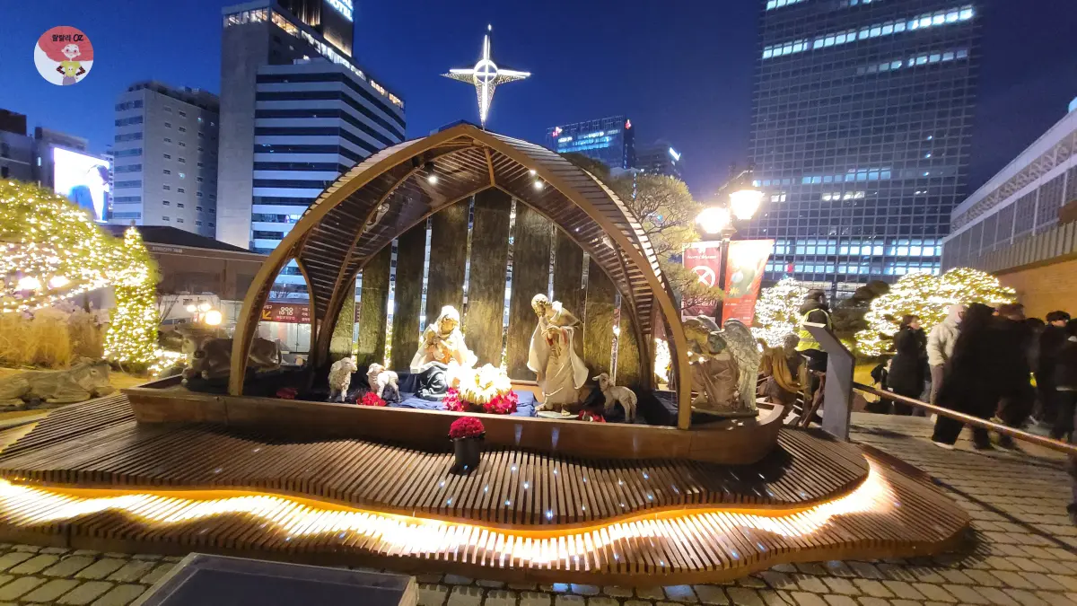 도심 야경 속 현대 건물들을 배경으로, 환하게 빛나는 명동성당의 야외 성탄 구유 전시 모습.
