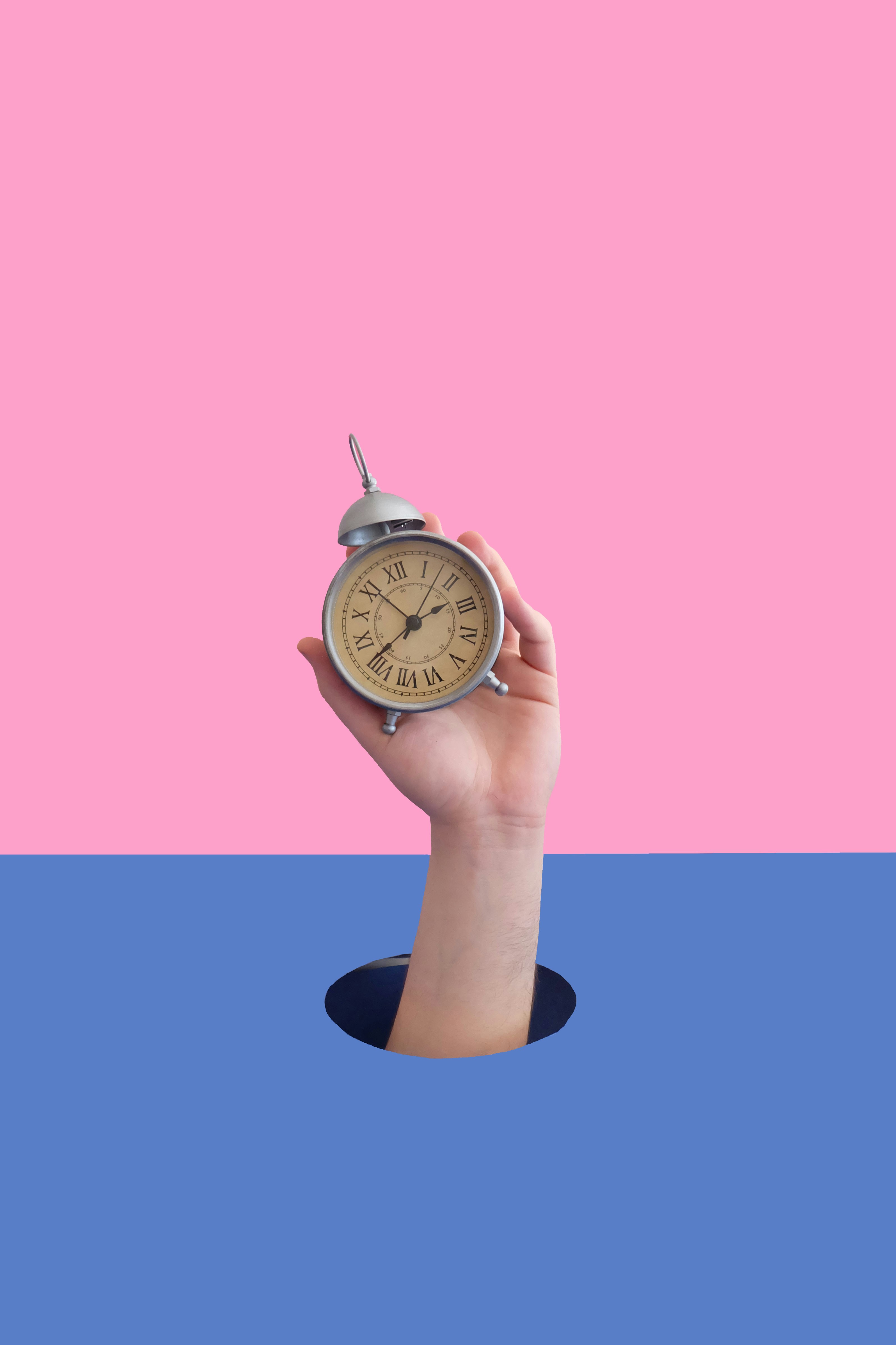 핑크와 파란 배경 위에서 손이 알람시계를 들고 있는 장면 – 시간 개념을 상징하는 이미지