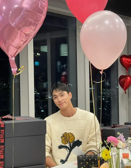 솔로지옥 3- 회색 선물상자가 많은 주변 분홍색, 빨강색 풍선 가운데 아이보리색 긴팔 티를 입고 의자에 앉아 미소짓고 있는 남자출연자 박민규