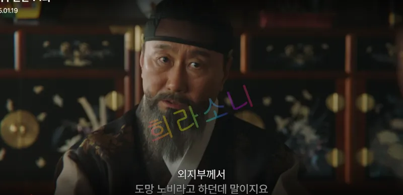 박준기가 화면 정면을 바라보며 진지한 표정을 짓고 있다. 그는 머리에 검은 두건을 쓰고 갈색 조끼와 흰 저고리를 입었다. 그의 뒤에는 장식이 있는 전통 목가구들이 놓여 있다. 자막: &ldquo;외지부께서 도망 노비라고 하던데 말이지요.&rdquo;