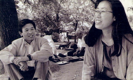 젊은 시절의 부부 가수 정태춘과 박은옥이 야외 공원에서 나란히 앉아 웃으며 대화하고 있는 흑백 사진. 뒤편에는 나무 그늘 아래 바둑을 두고 있는 노인들이 보인다.