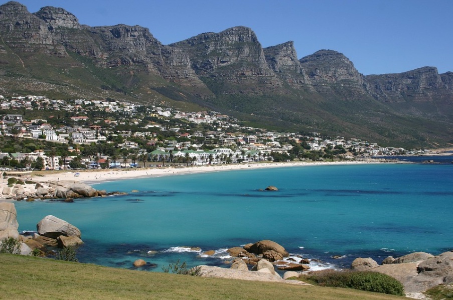 남아프리카 공화국 케이프타운의 캠스 베이(Camps Bay) 해안가 전경. 화면 상단에는 웅장한 '열두 사도 봉우리(Twelve Apostles)' 산맥이 펼쳐져 있고, 그 아래로 에메랄드빛 바다와 하얀 모래사장, 그리고 해안선을 따라 형성된 평화로운 마을 모습.