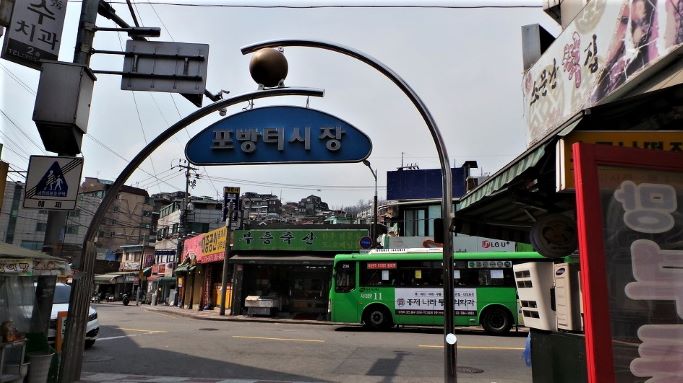 포방터 시장 입구, 원형 철봉 이중 조형물, 녹색 마을버스, 여러 간판들,