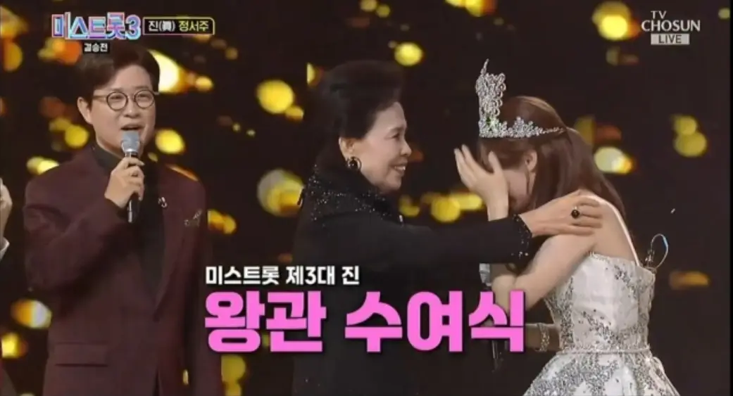 미스트롯3-
검은색 금빛 꽃이 내리는 배경 앞 좌측 붉은색 쟈켓을 입은 진행자가 마이크를 들고 말하고 중앙 검은옷을 입은 이미자가 우승한 정서주의 어깨를 만져주며 웃늠 모습 앞 흰글씨 미스트롯 제 3대 진
아래 분홍글씨 왕관수여식