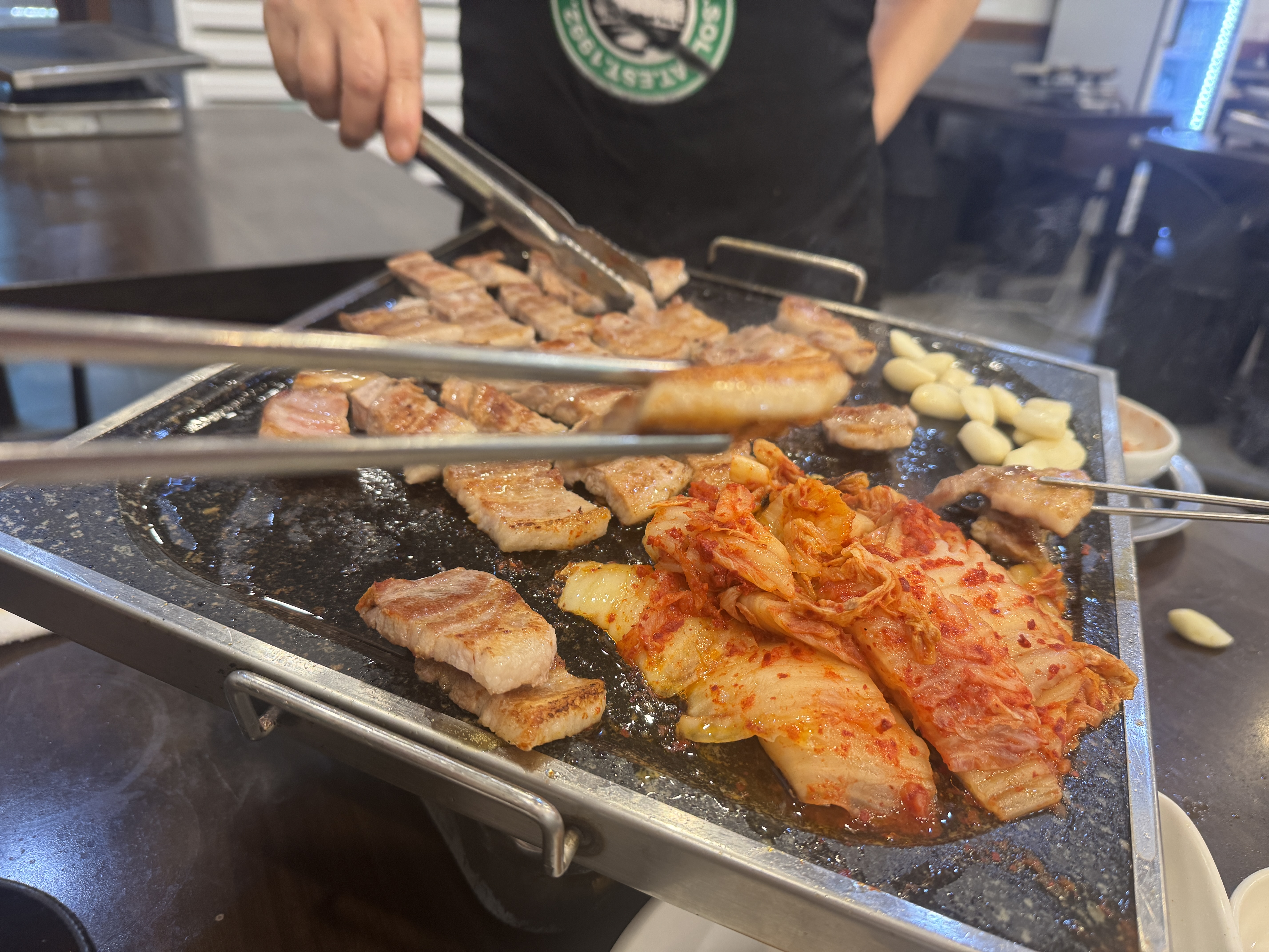 한 조각의 삼겹살을 젓가락으로 집어드는 장면. 불판 위 고기들이 노릇하게 익어가고 있다.