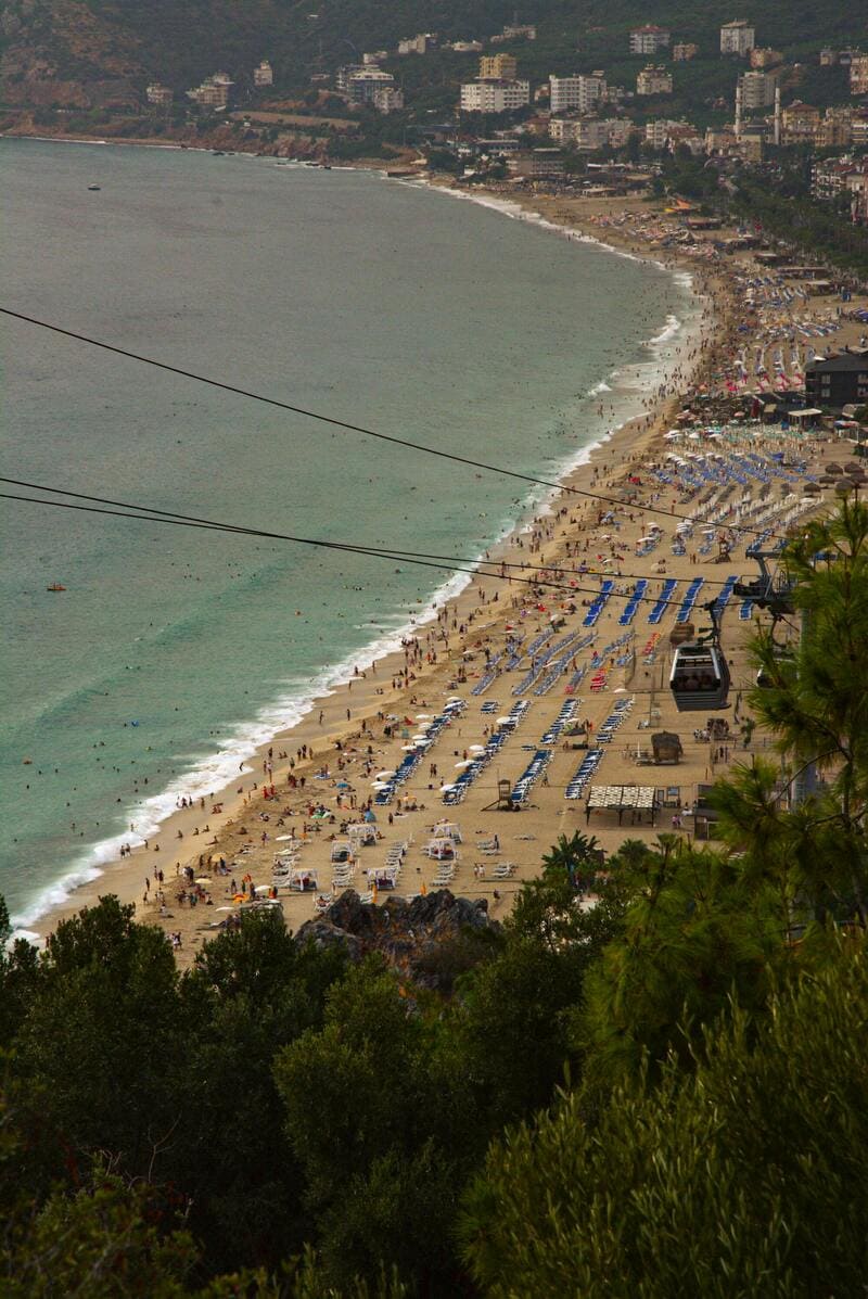 안탈리아 해안의 대형 해변 풍경. 모래사장에는 파라솔과 썬베드가 줄지어 있고, 사람들로 북적이는 모습이 한눈에 들어옵니다. 오른쪽 상단에는 해변을 따라 이동하는 케이블카가 보입니다