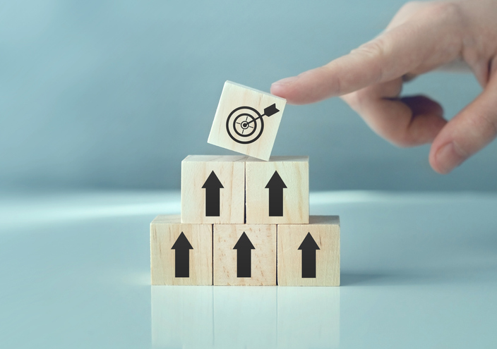 목표 아이콘이 그려진 나무 블록을 위로 쌓아 올리며 단계적으로 목표 달성을 상징하는 이미지