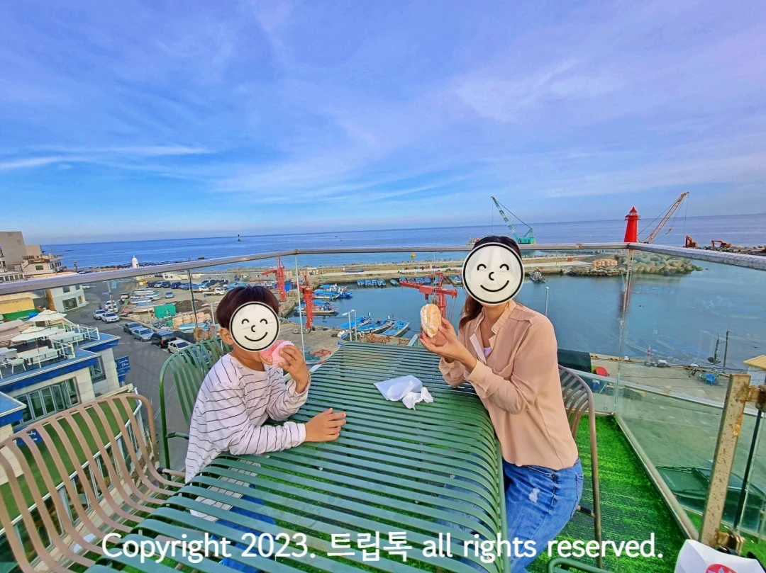 넓은 하늘과 방파제와 빨간 등대와 바다를 배경으로 두사람이 테이블에 앉아 도넛을 먹고 있다